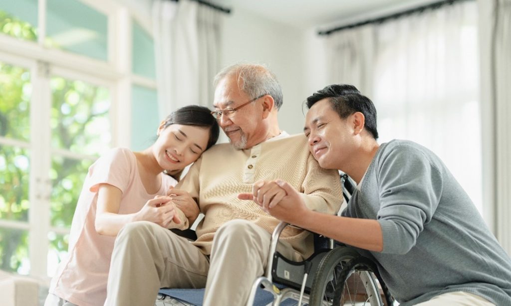 Disabled man in wheelchair with two young adults either side hugging him