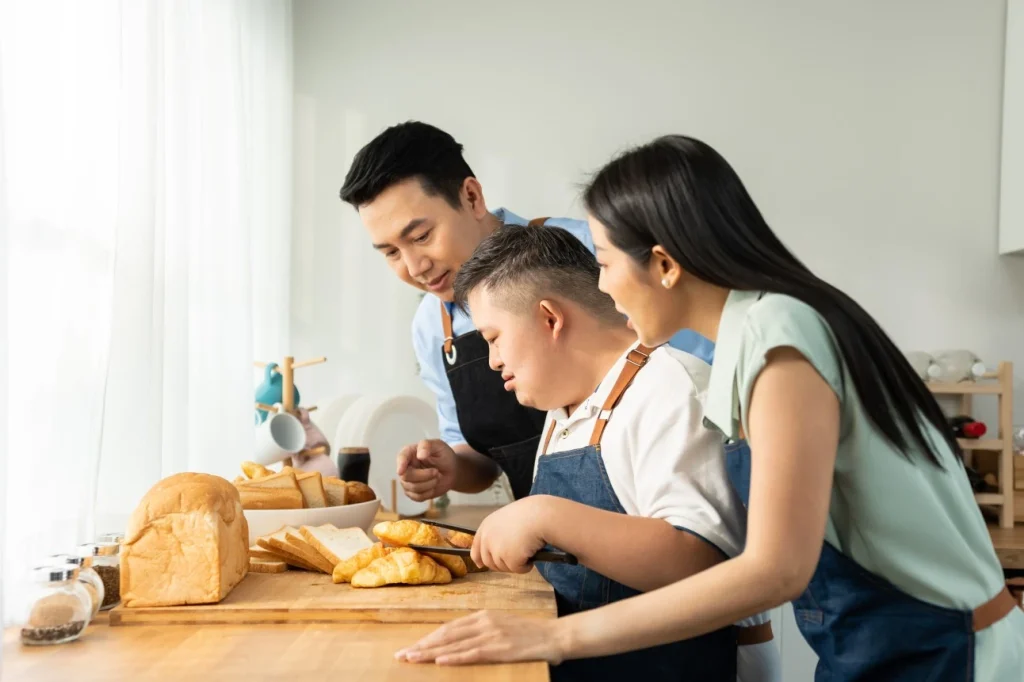 Disabled man using tongs to pick up croissant in kitchen with two support workers